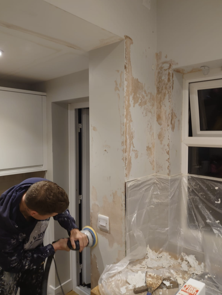A person using a sander on a wall with damaged, peeling paint near a window in a kitchen.