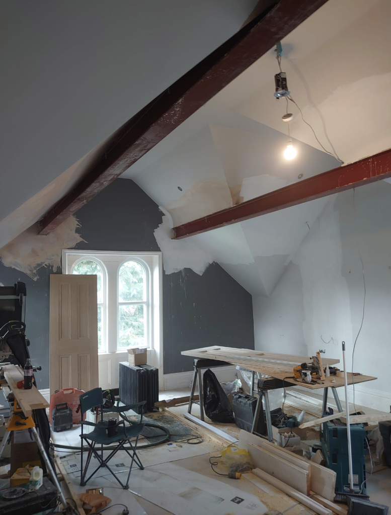 An attic room under renovation, featuring exposed steel beams, walls partially painted grey, and various construction tools.
