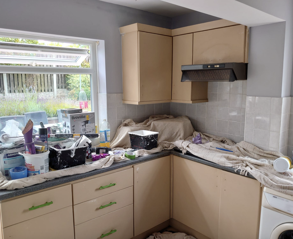 A kitchen undergoing renovation with beige cabinets, grey walls, and painting supplies spread across the countertops.