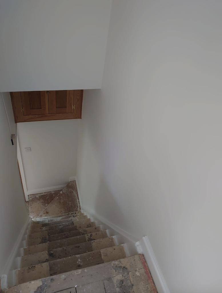 A steep, narrow staircase under construction with bare, unfinished steps, white walls, and a small wooden cabinet above.