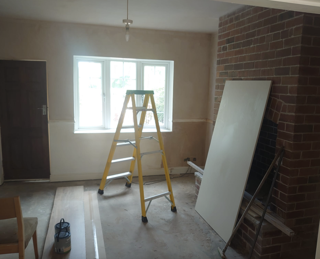 A room under renovation featuring a yellow stepladder, a white board leaning against a brick fireplace, and bare floors.