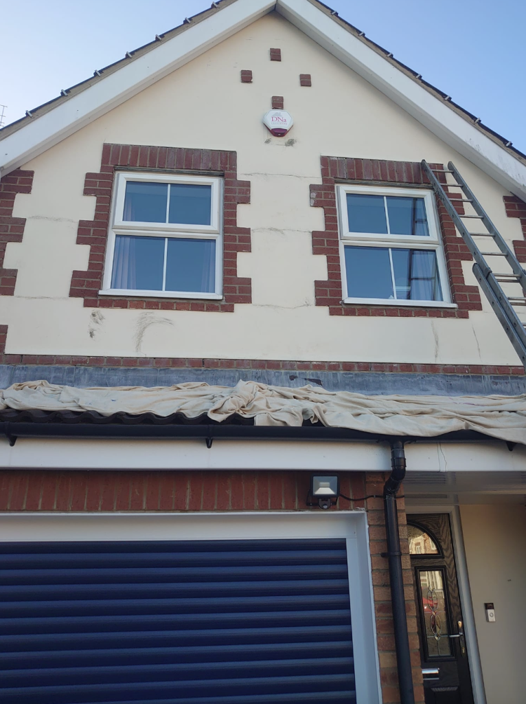 A residential house front with two upper-story windows, brick detailing, a garage, and a ladder leaning against the wall.