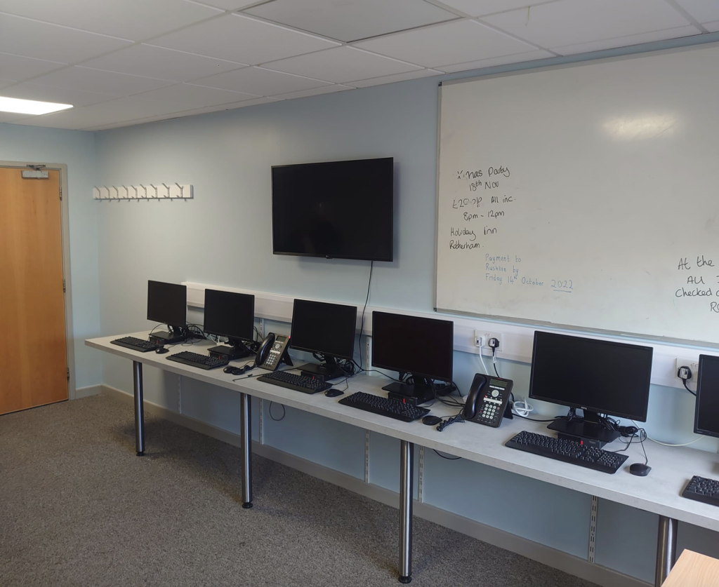 A row of computer workstations arranged along a wall in an office, with a wall-mounted TV and a whiteboard.