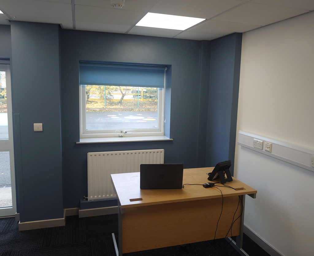 A modern office space with a desk, laptop, and phone, featuring dark blue walls, a window with a blind, and white trim.