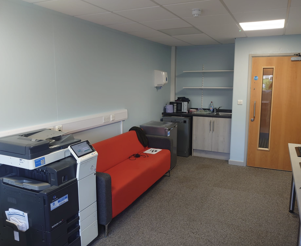 A small office space featuring a large printer, an orange sofa against a light blue wall, and a kitchenette in the corner.
