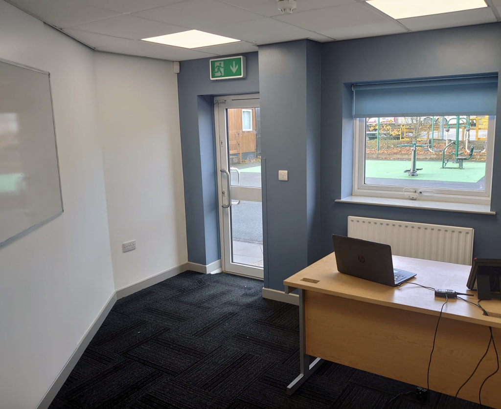 A bright office with dark carpet, a desk with a laptop, a glass door, and a large window looking out to a playground.