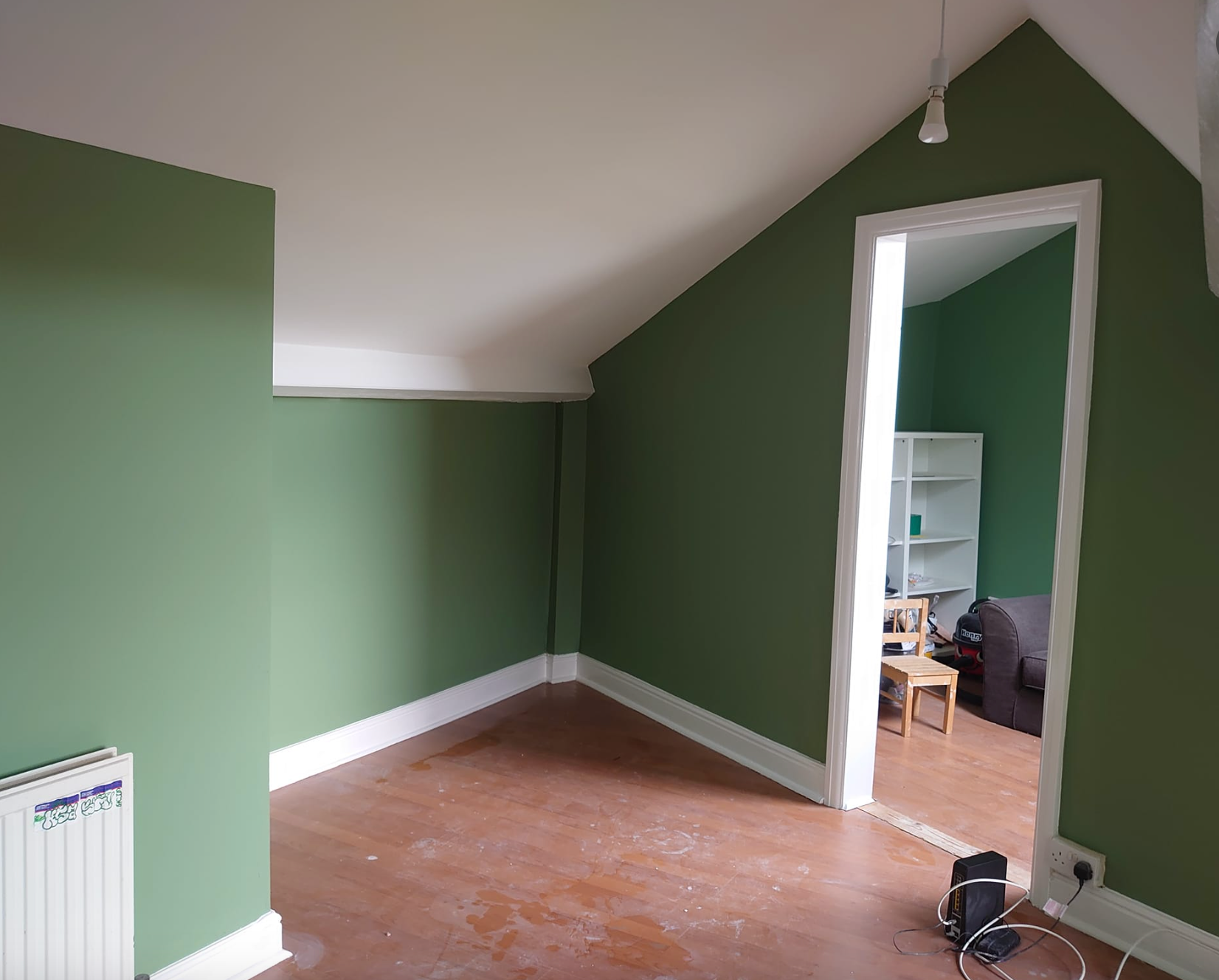 An attic room with green walls and white trim, featuring a slanted ceiling, a heater, and a doorway to another room.