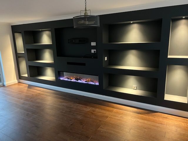 Modern dark-charcoal custom wall unit featuring built-in shelving, a recessed electric fireplace, and warm accent lighting.