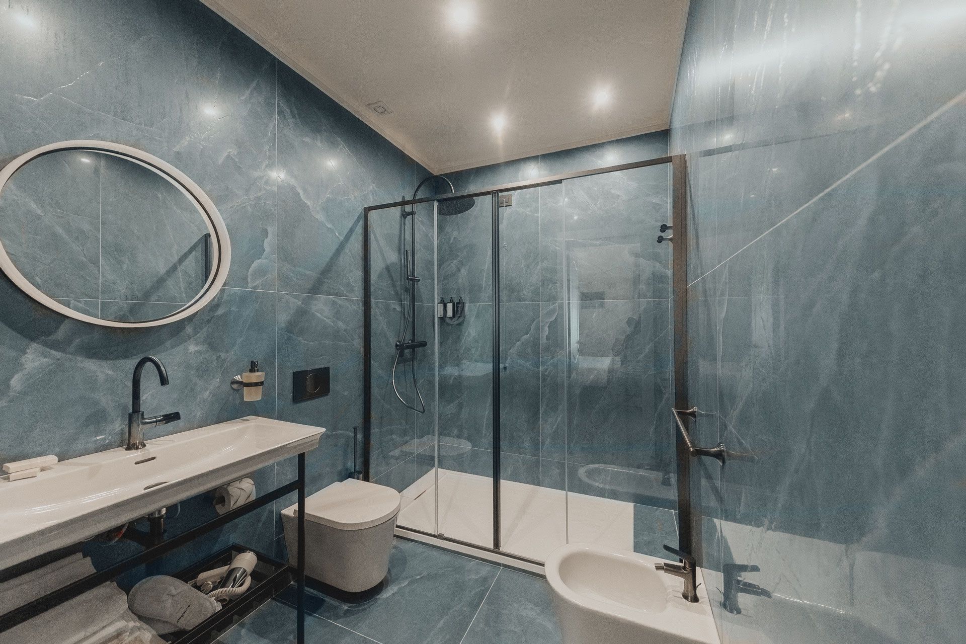 Bagno moderno con pareti in marmo blu, doccia in vetro, lavabo con mobiletto, WC e specchio rotondo.