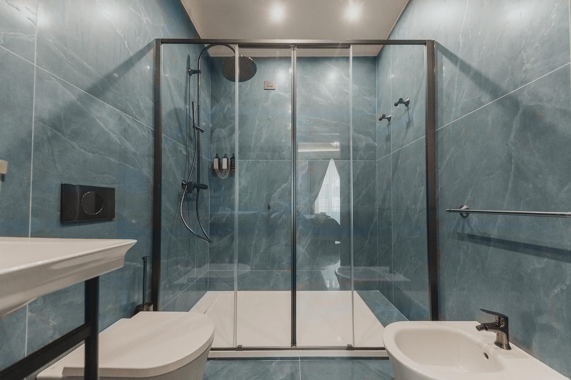 Bagno moderno con piastrelle blu, doccia in vetro, lavabo bianco, WC e specchio per il trucco.