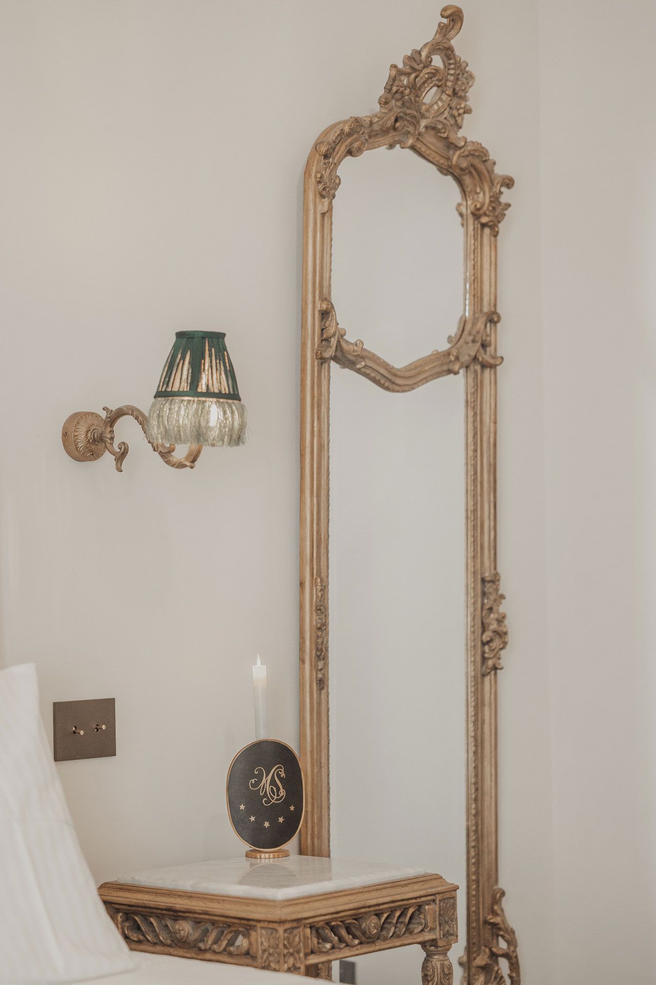 Specchio decorato con cornice dorata sopra un tavolino ornamentale accanto a un'applique in una stanza dai colori chiari.