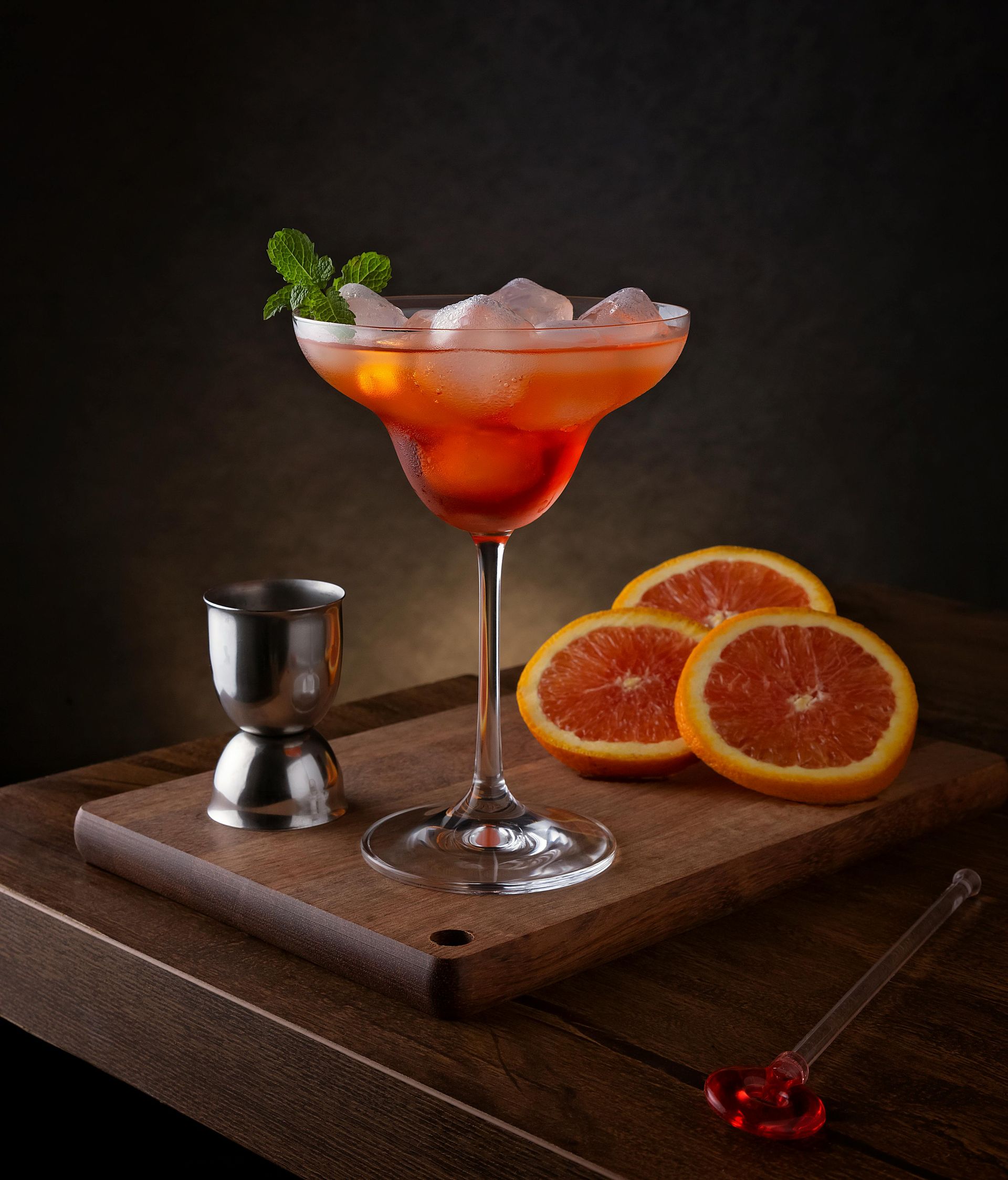 Cocktail all'arancia in un bicchiere da martini con ghiaccio, guarnizione di menta e fette di agrumi su un tagliere di legno.
