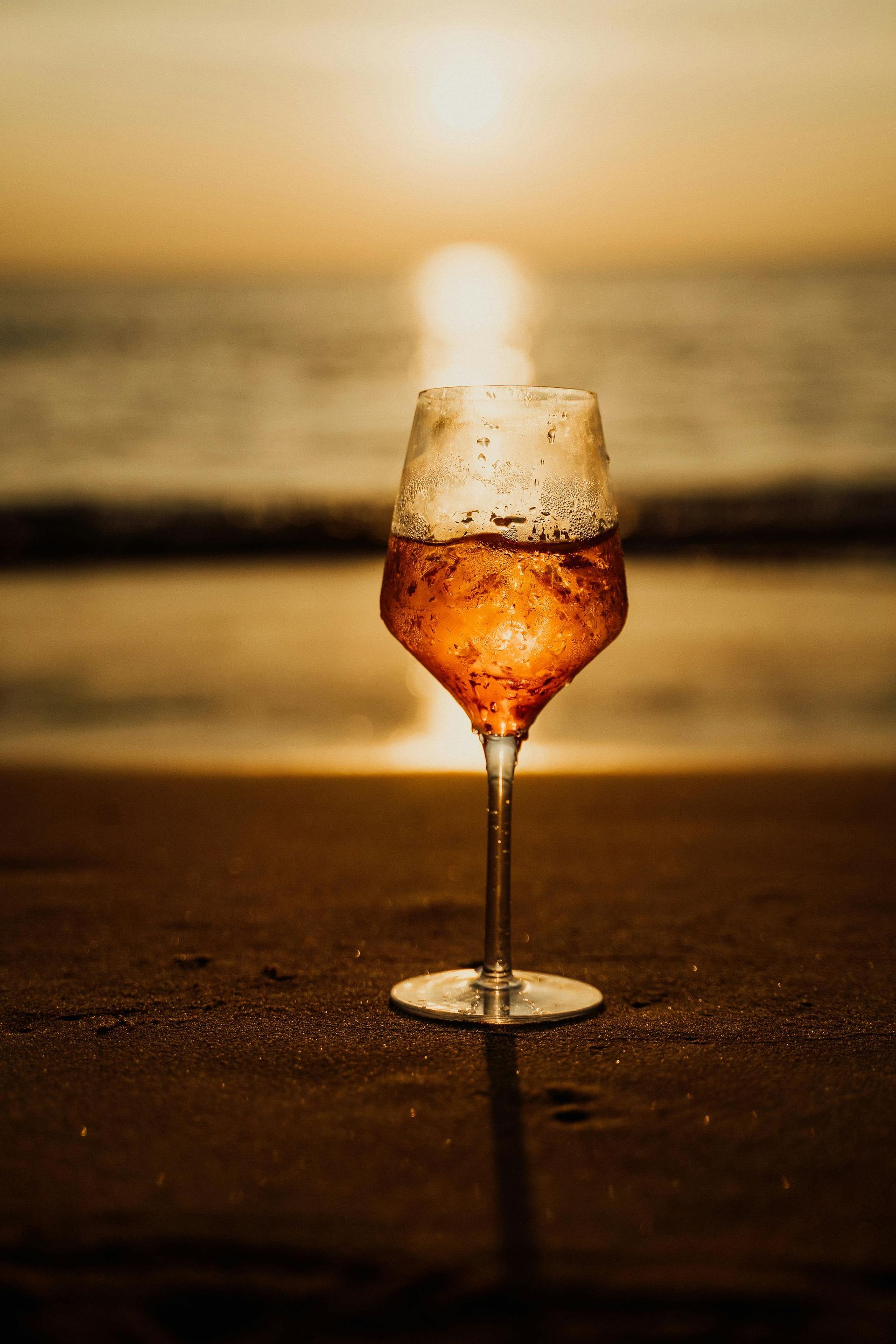 Un calice di vino su una spiaggia bagnata al tramonto, con la luce dorata che si riflette sulla sabbia e sul mare.