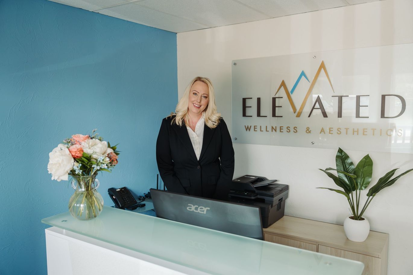 A woman is standing at a desk in front of a sign that says elevated wellness & aesthetics.
