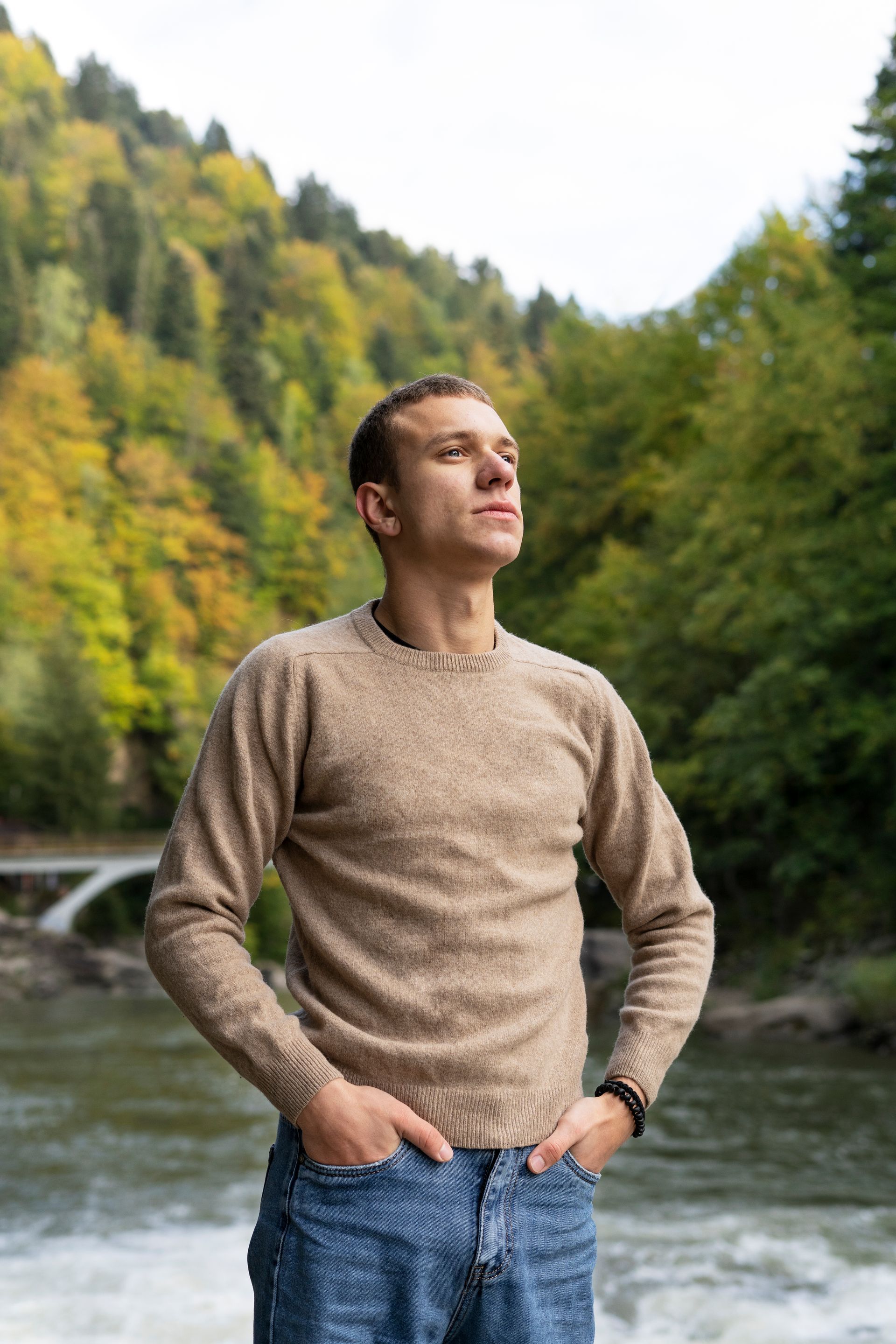 A man in a sweater is standing in front of a river with his hands in his pockets.