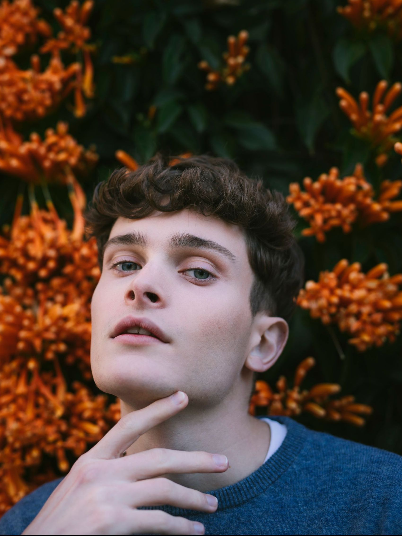 A young man in a blue sweater is standing in front of flowers.