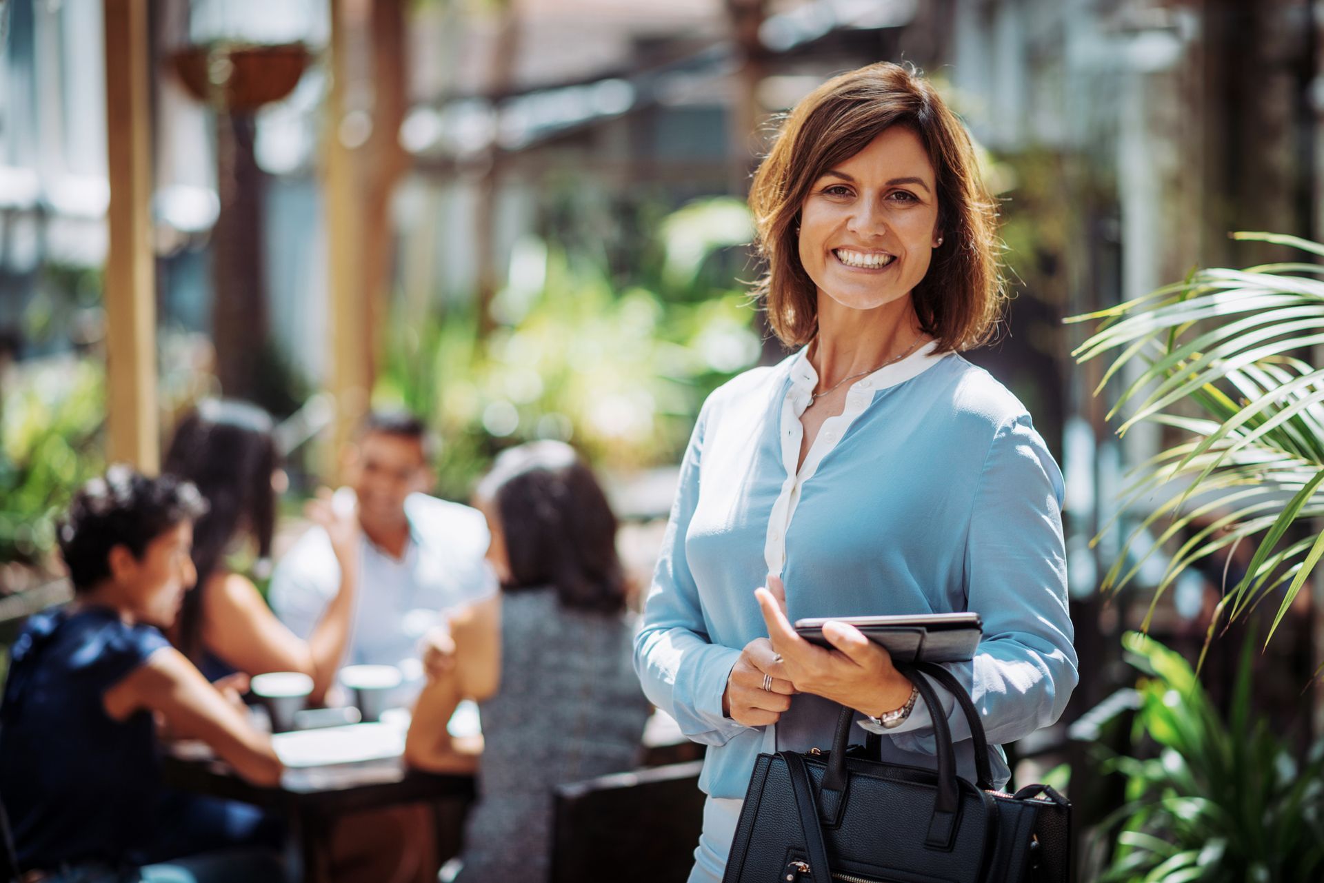 A woman is standing in front of a restaurant holding a tablet and a purse.