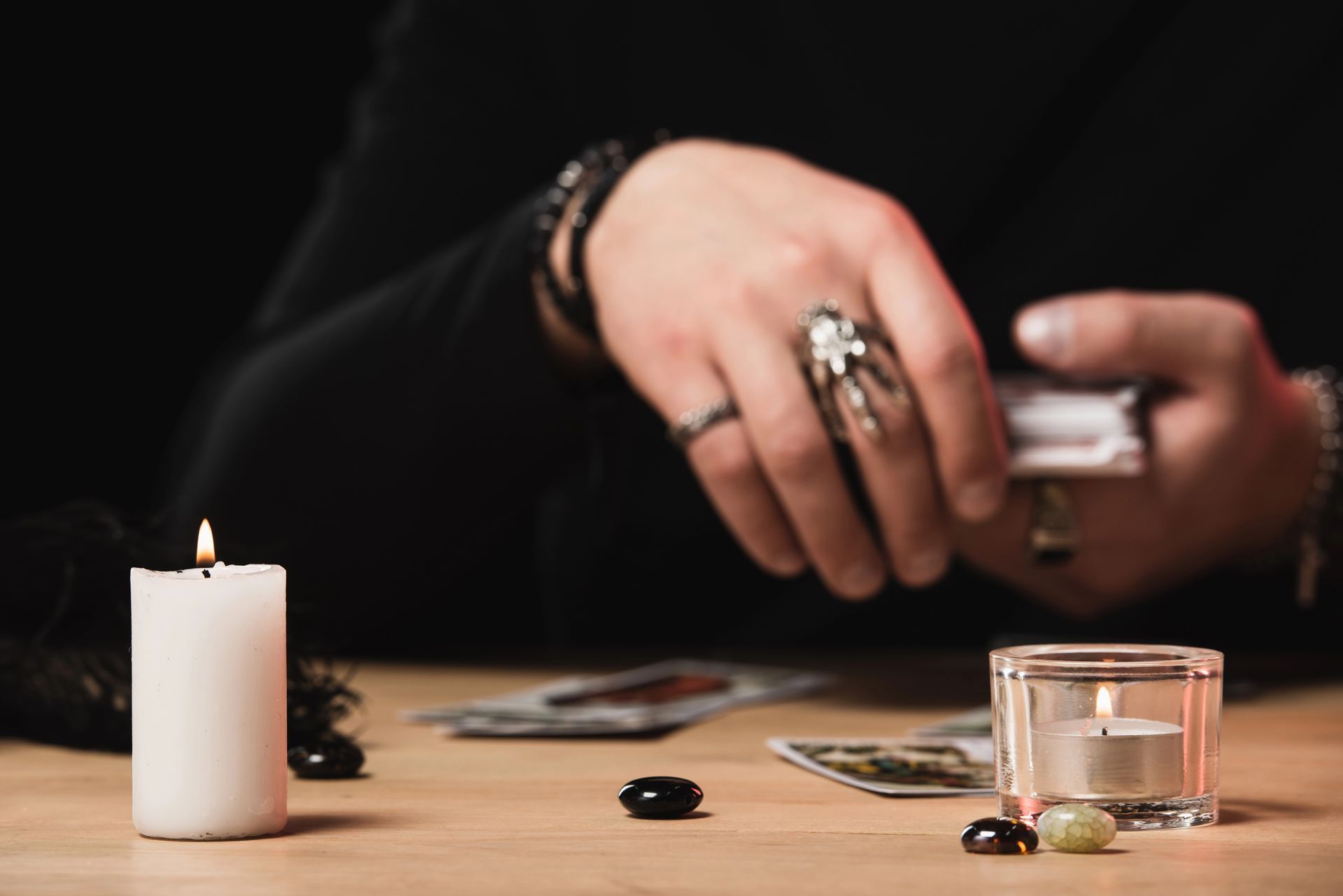 Manos con anillos y pulseras manipulan cartas junto a una vela encendida y un vaso sobre una mesa oscura.