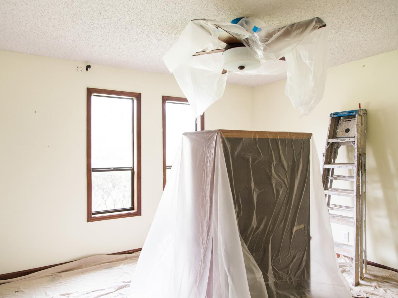 Room prepared for renovation, furniture and fan covered in plastic. Ladder stands nearby.