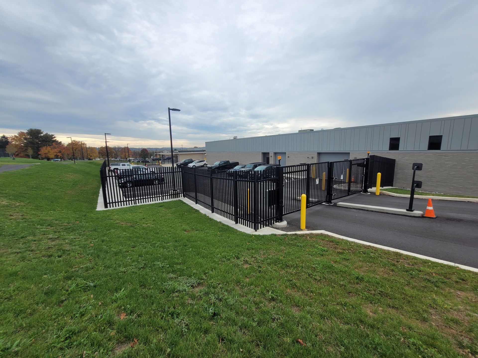 Black metal barricades around a sidewalk beside a gray building, with a grassy field and cloudy sky.