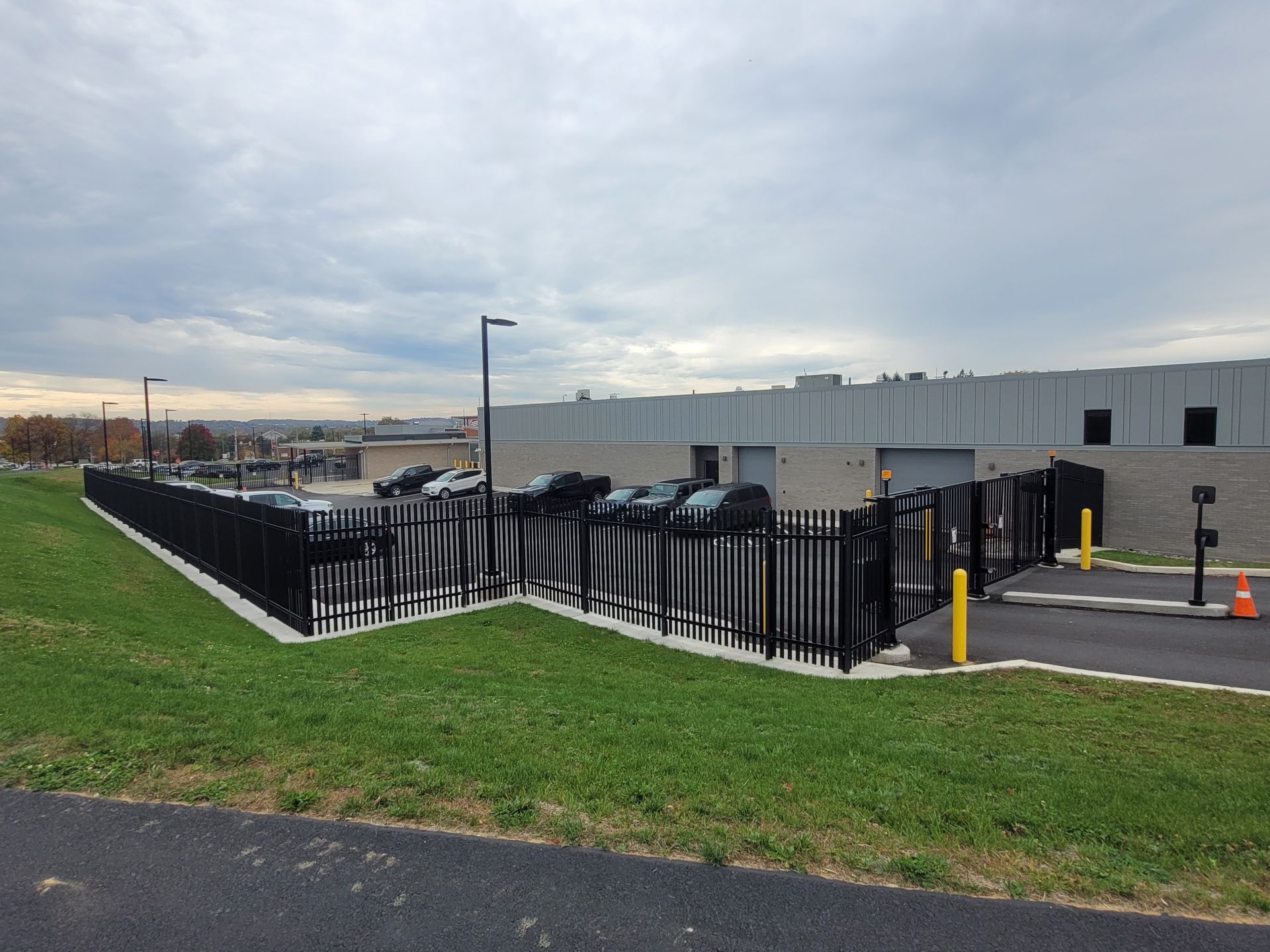 Black metal fencing around a utility facility beside a grassy verge under a cloudy sky