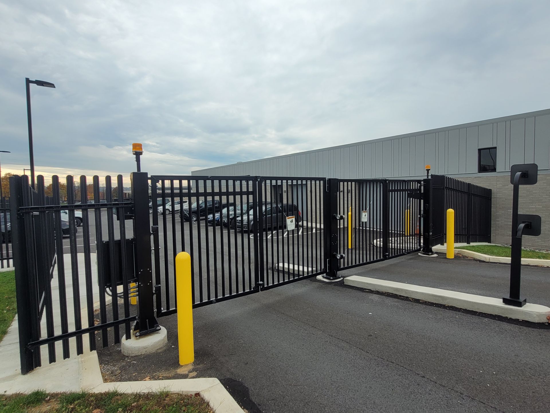 Black metal gate at a warehouse entrance with yellow bollards and cloudy sky.