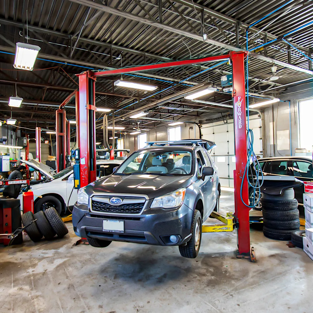 A gray Subaru Forester on a lift in a car repair shop; other vehicles and tires visible.