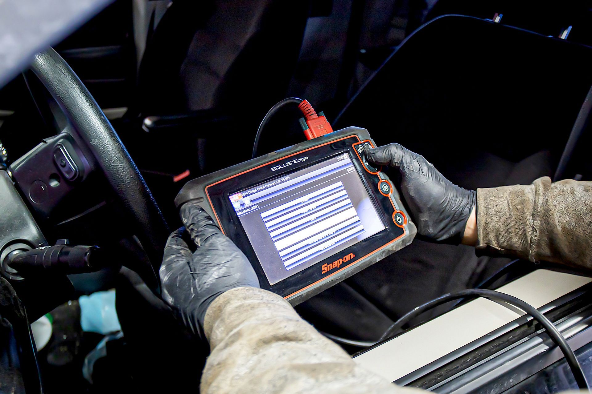 A person in black gloves using a diagnostic scanner inside a car. The device shows multiple lines of data.