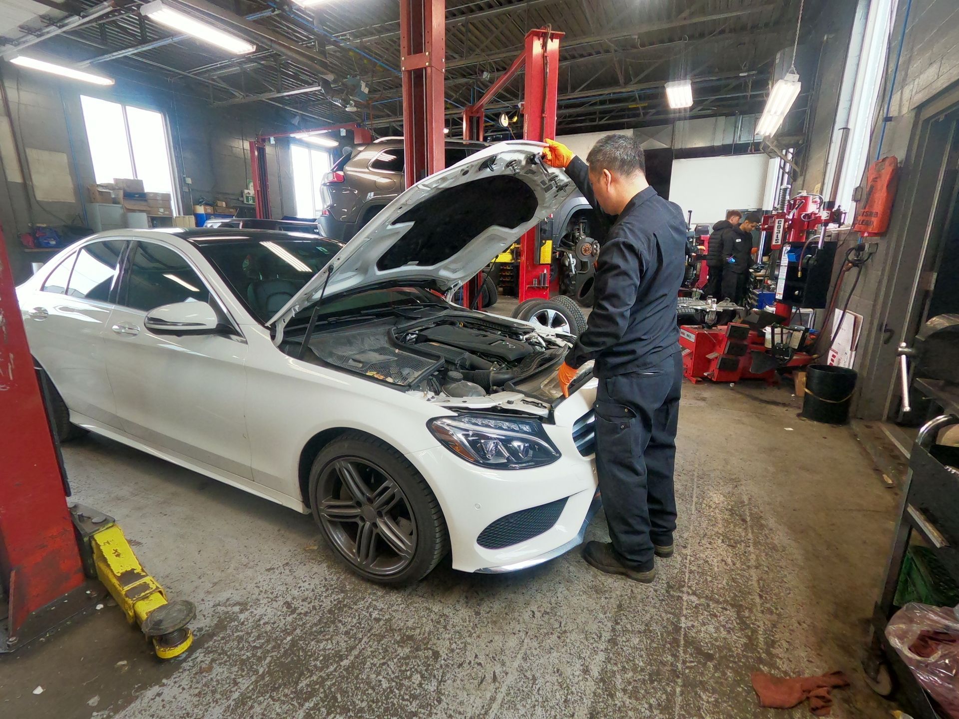Mechanic in blue jumpsuit works on white car with open hood in a garage.