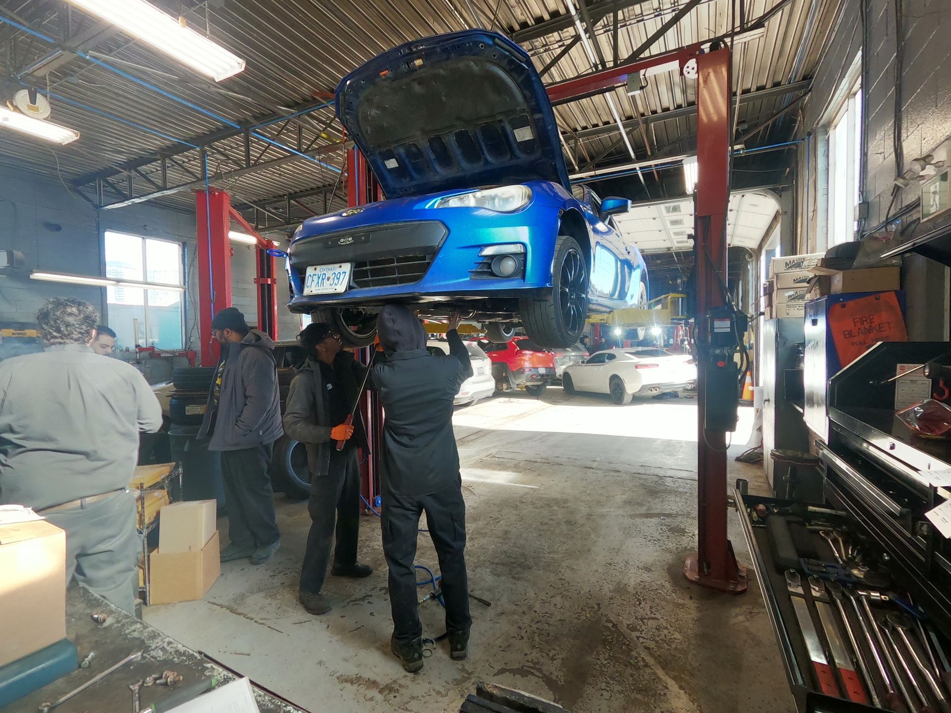 Blue car on a lift in a repair shop, mechanics working underneath, open hood.