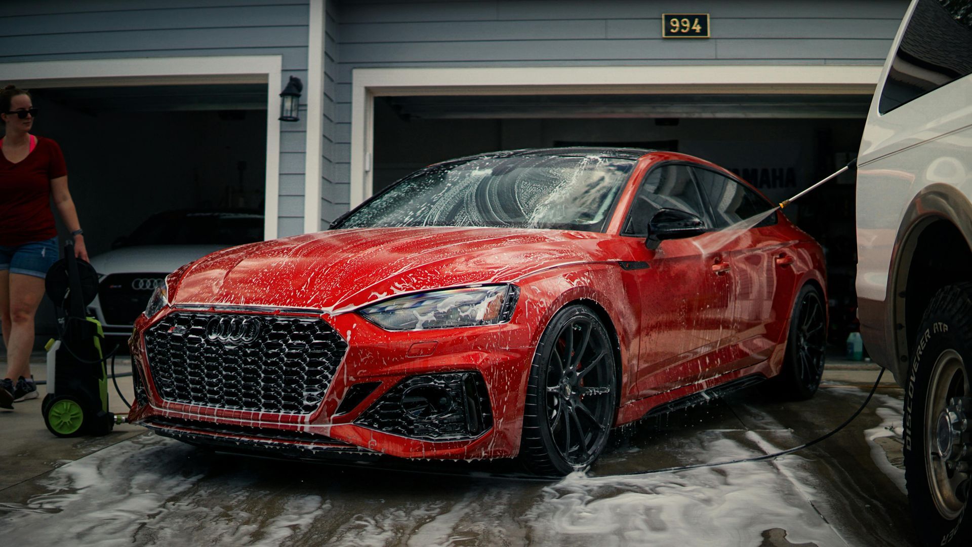 Red sports coupe being washed in a driveway, with soap suds and garage in the background.