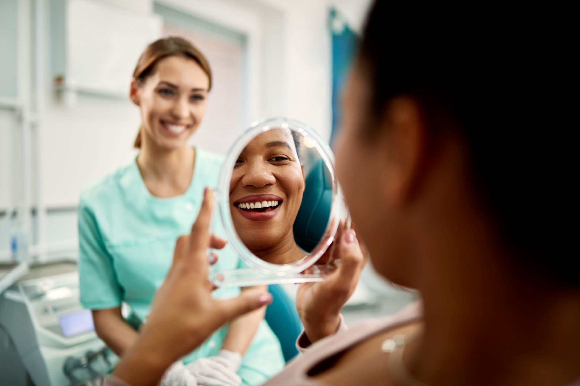 Woman smiles in a mirror while another person assists her in a bright room