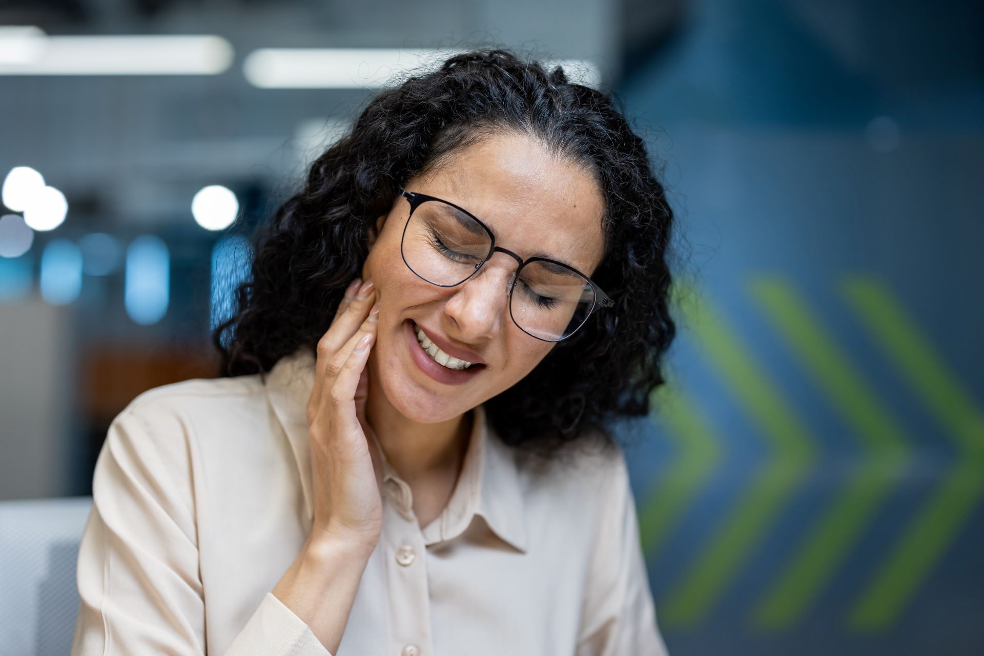 Woman with glasses holding her jaw, showing signs of pain in an office setting.
