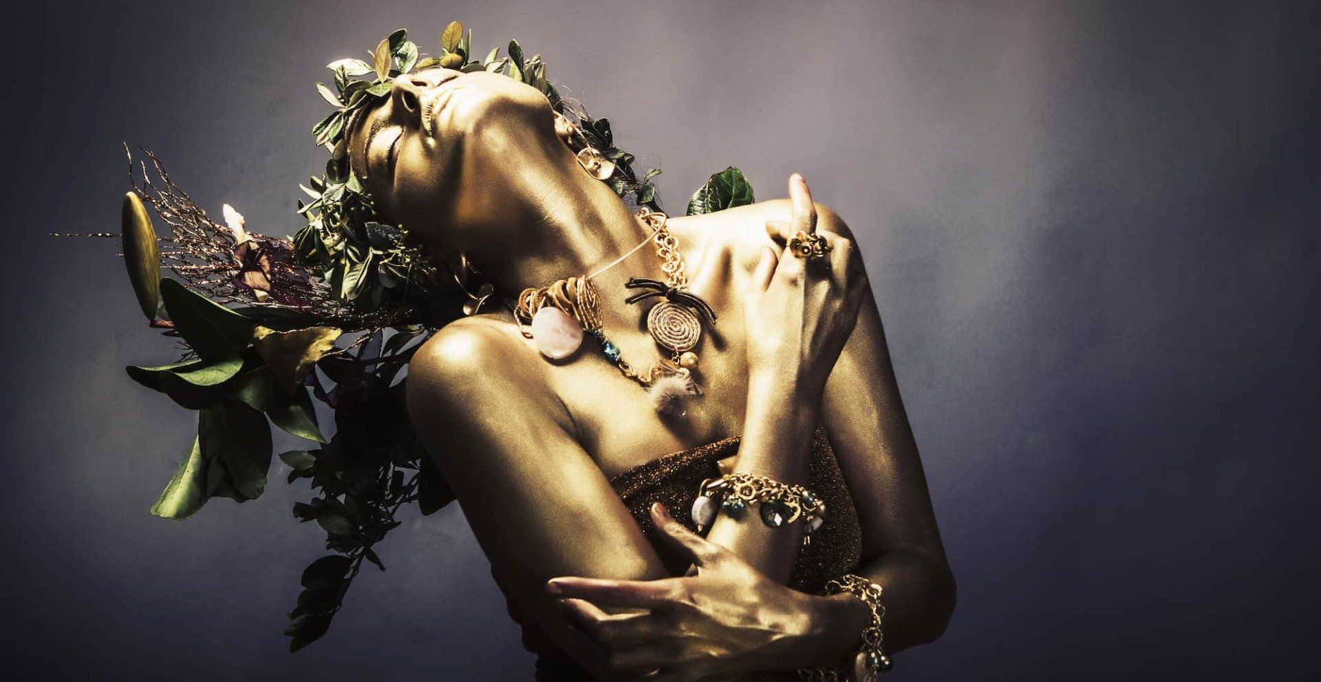 Gold-painted figure with closed eyes, head tilted back, and leafy adornments against a dark background