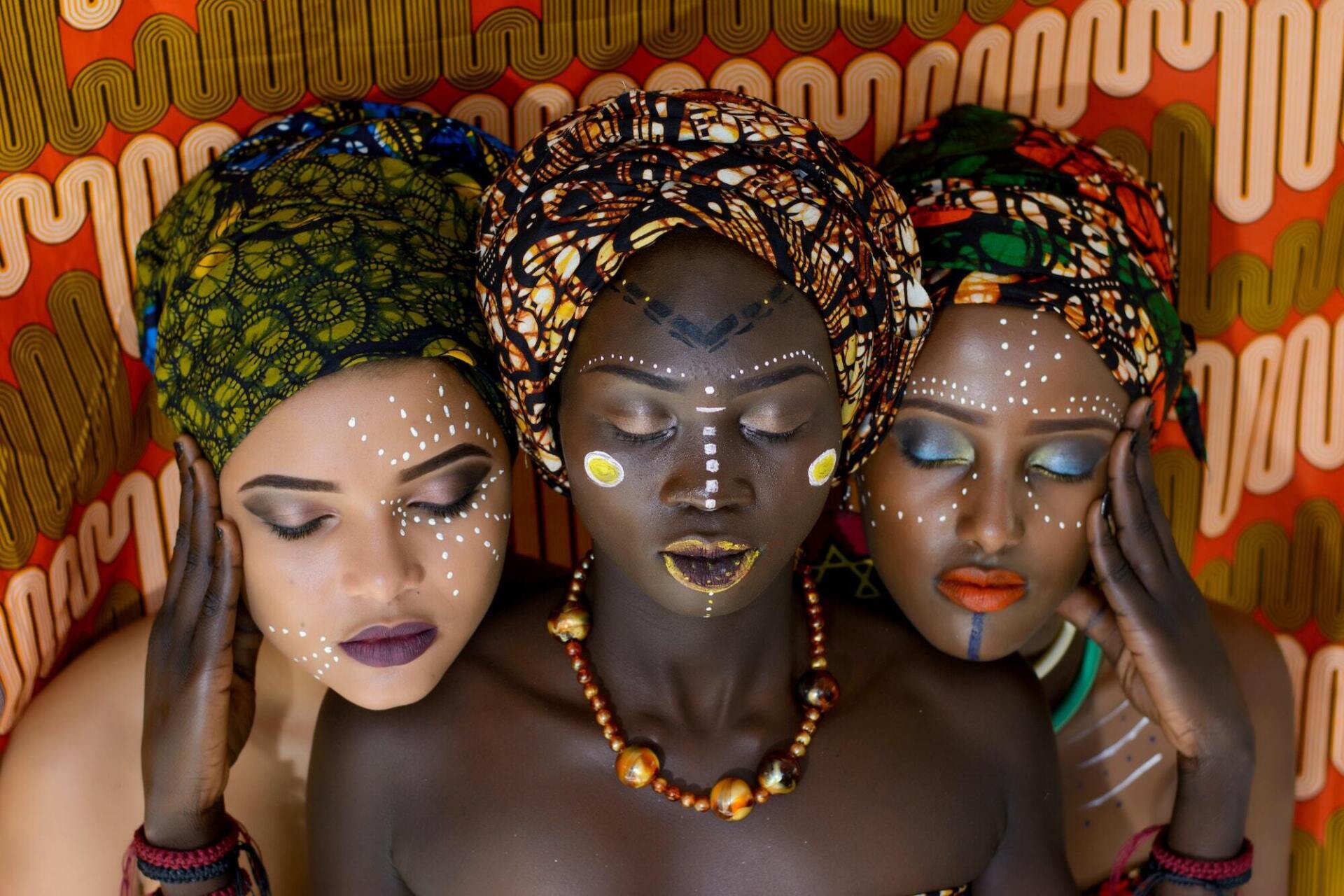 Three women with intricate headwraps and makeup pose against a patterned red and orange backdrop.