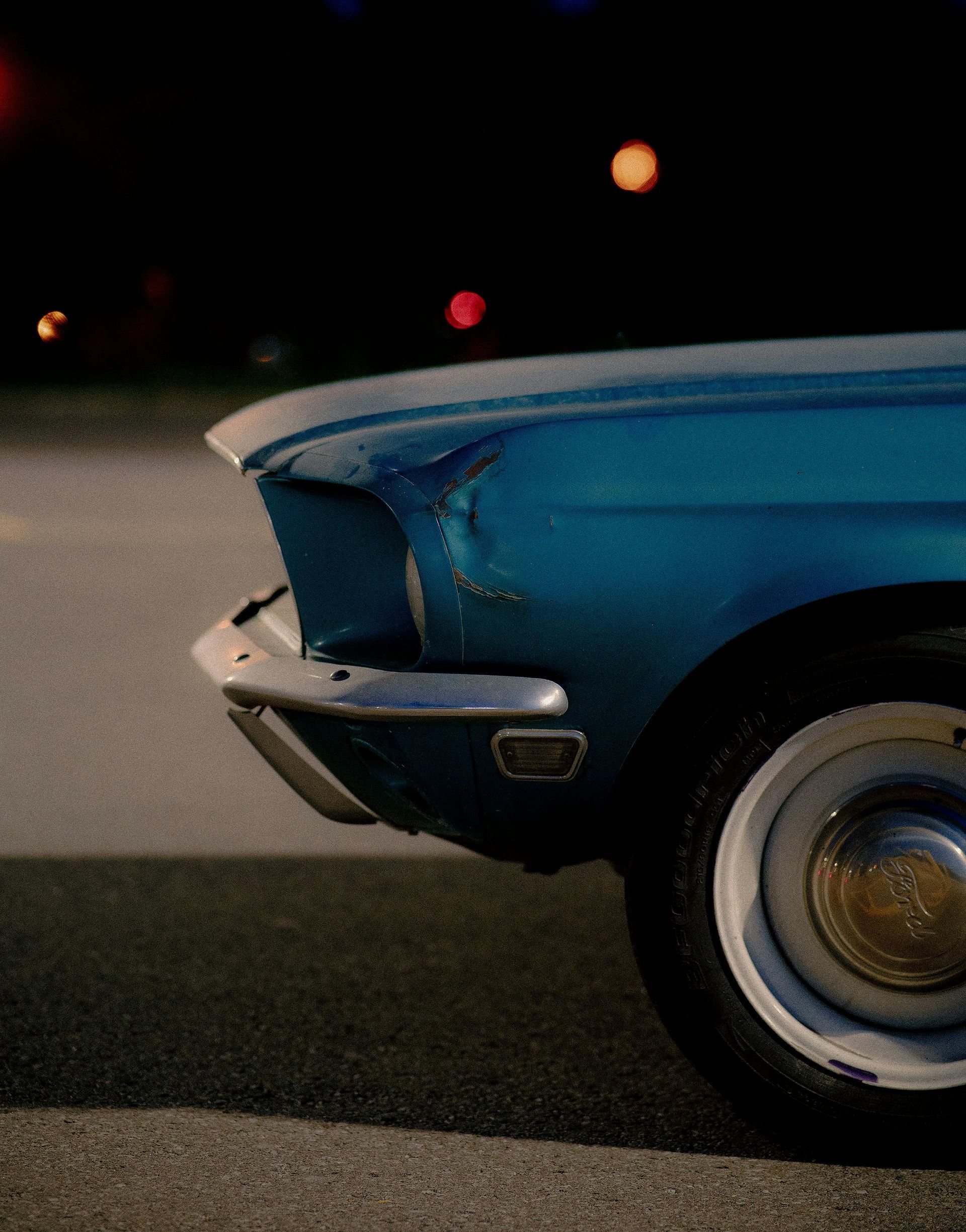 Blue classic car, partially visible, parked on pavement at night with blurred lights in the background.