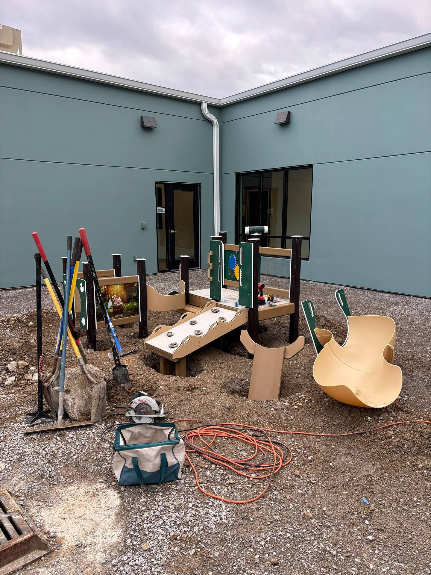 Construction tools and debris in a gravel courtyard beside a teal building wall