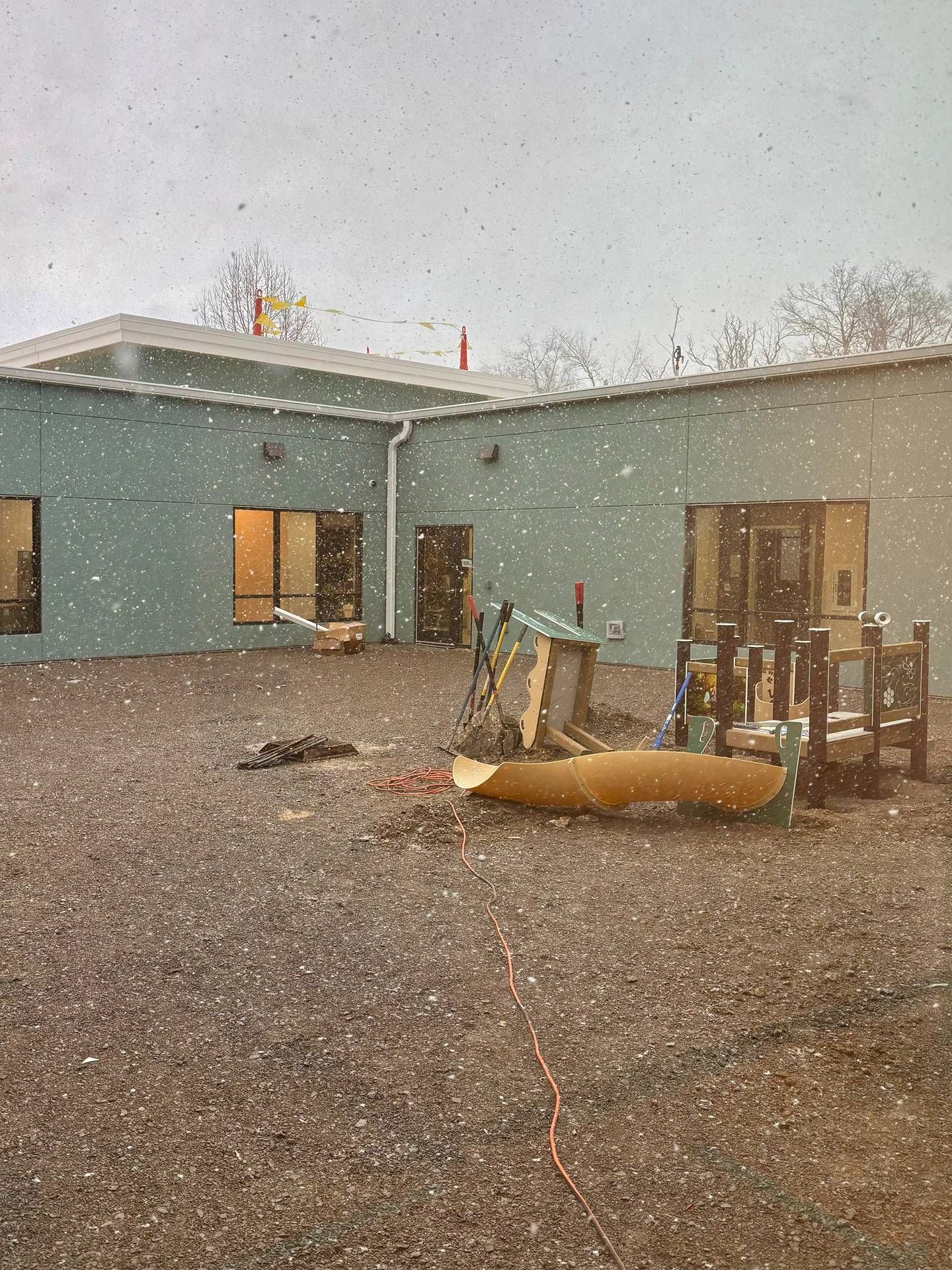 Snowy courtyard with a teal building, lit windows, and scattered construction materials.