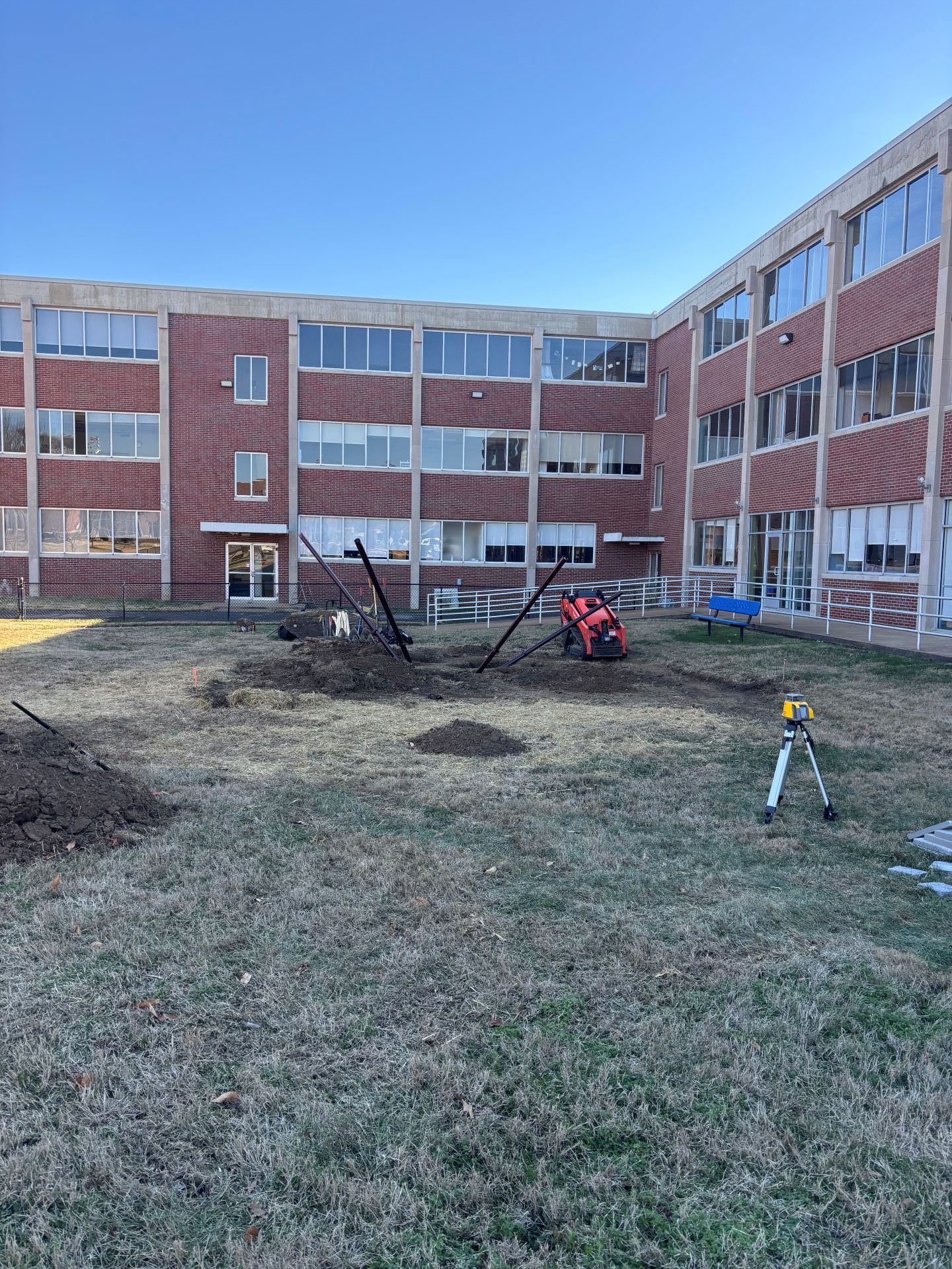 Courtyard of a brick school building with muddy ground and small playground equipment under a clear blue sky