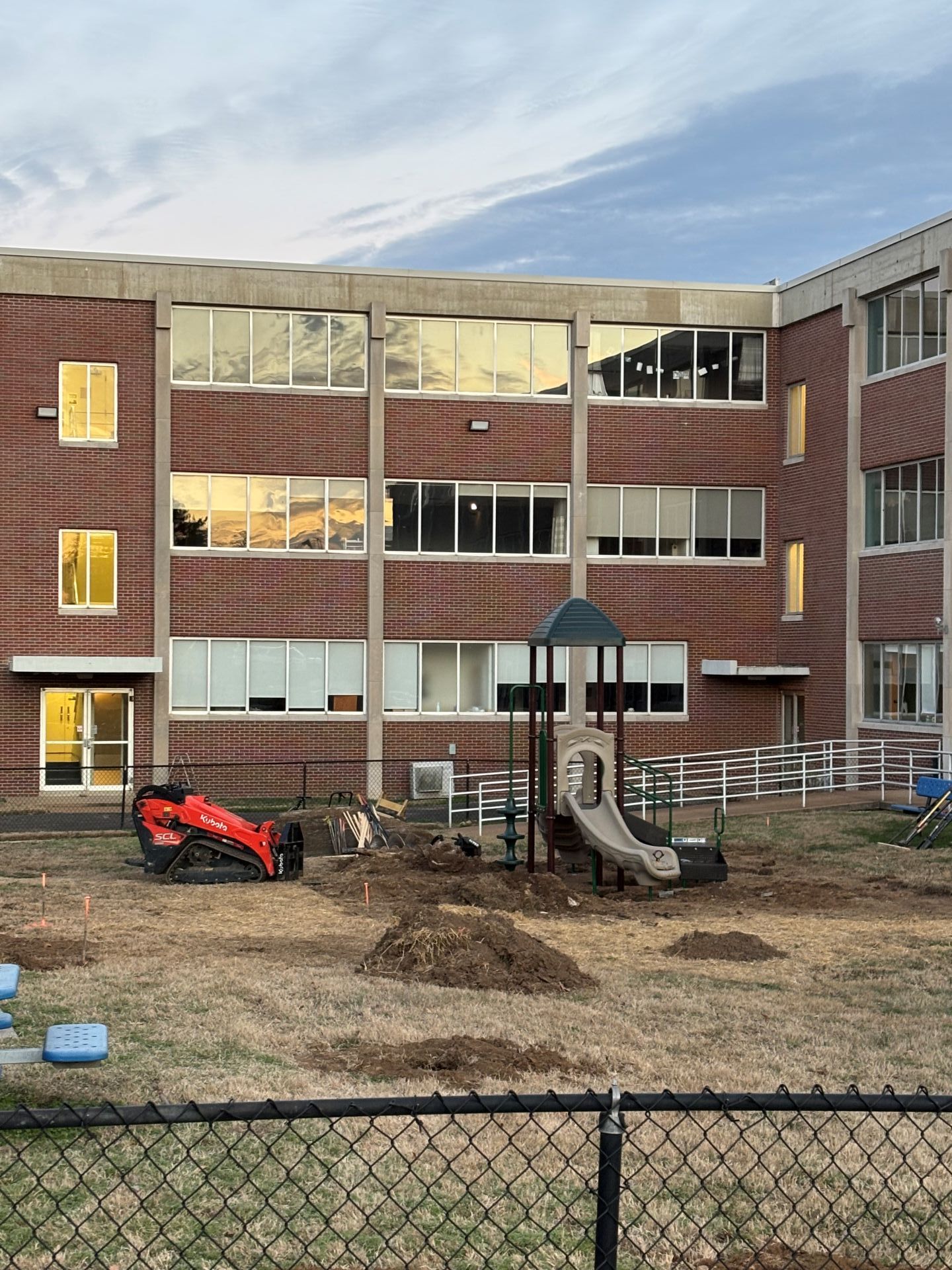 Playground with slide and digger toy in a fenced courtyard behind a brick apartment building