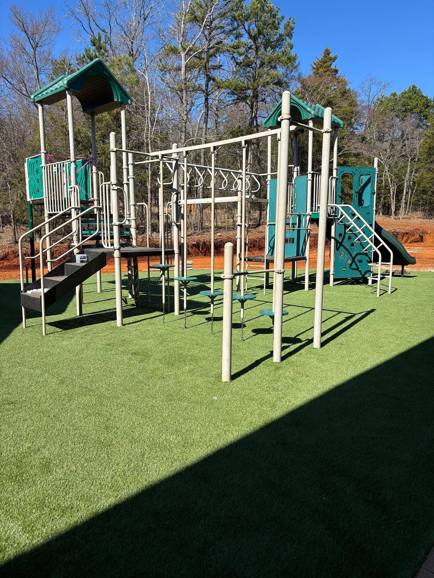 Outdoor playground with green turf, climbing bars, slides, and a shaded play tower under blue sky