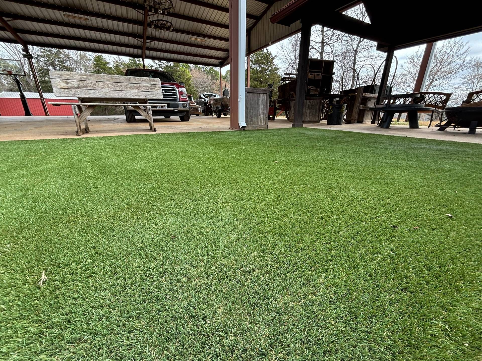 Covered outdoor seating area with green turf, picnic tables, and trees in the background