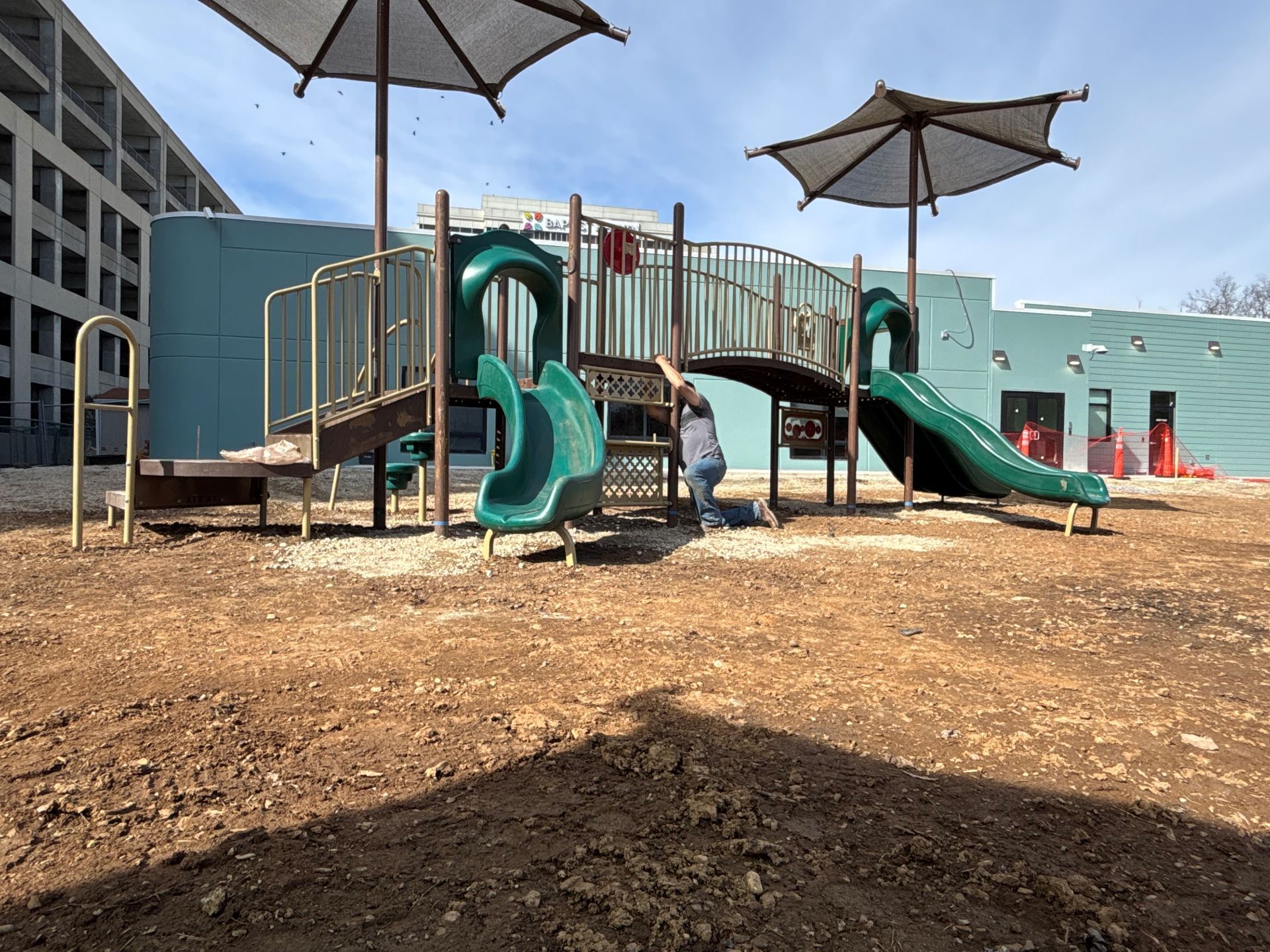 Outdoor playground with green slides, wooden bridge, and large shade umbrellas on a dirt surface