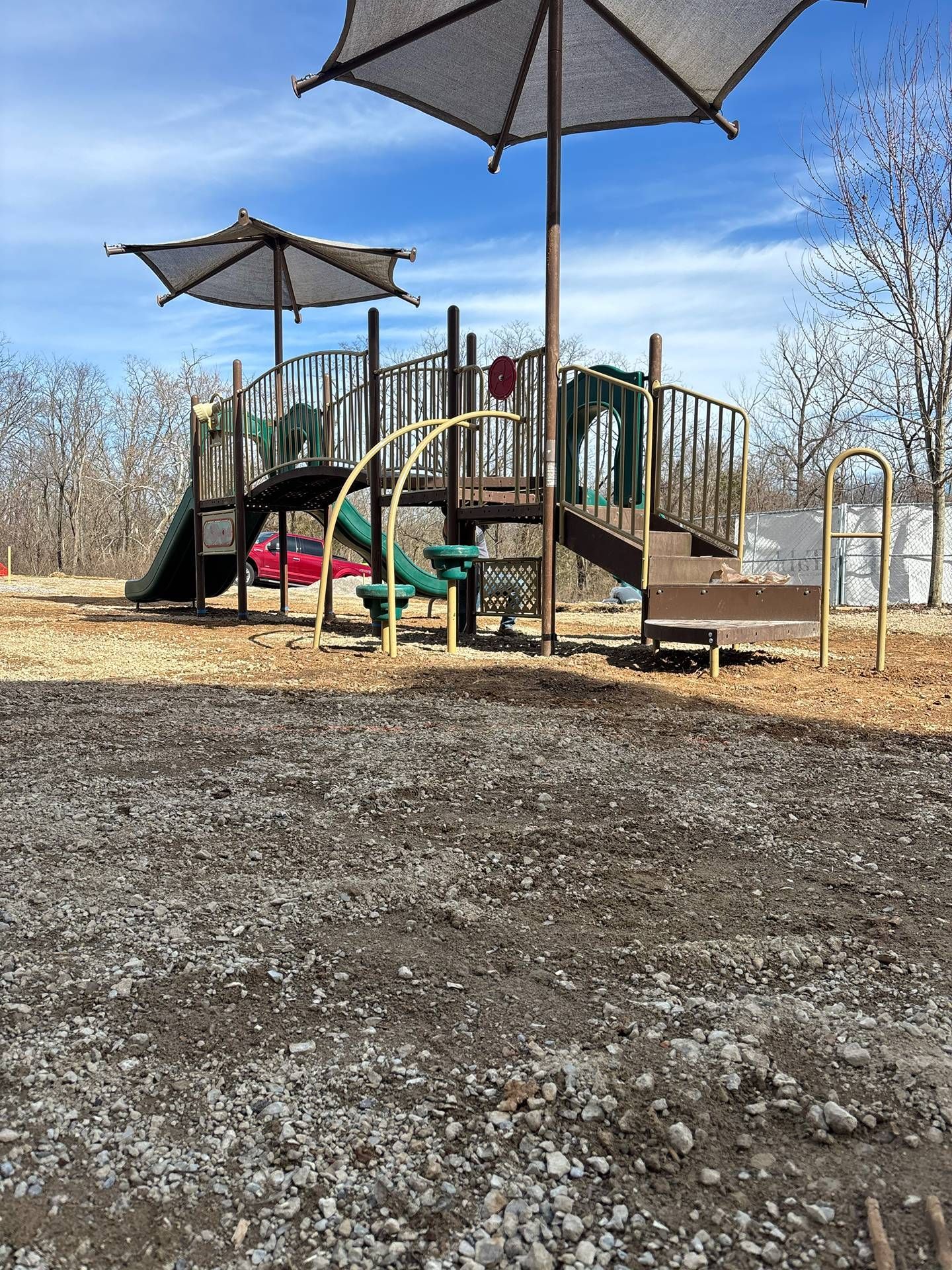Playground with climbing structures and sun umbrellas on a sunny day, surrounded by bare trees.