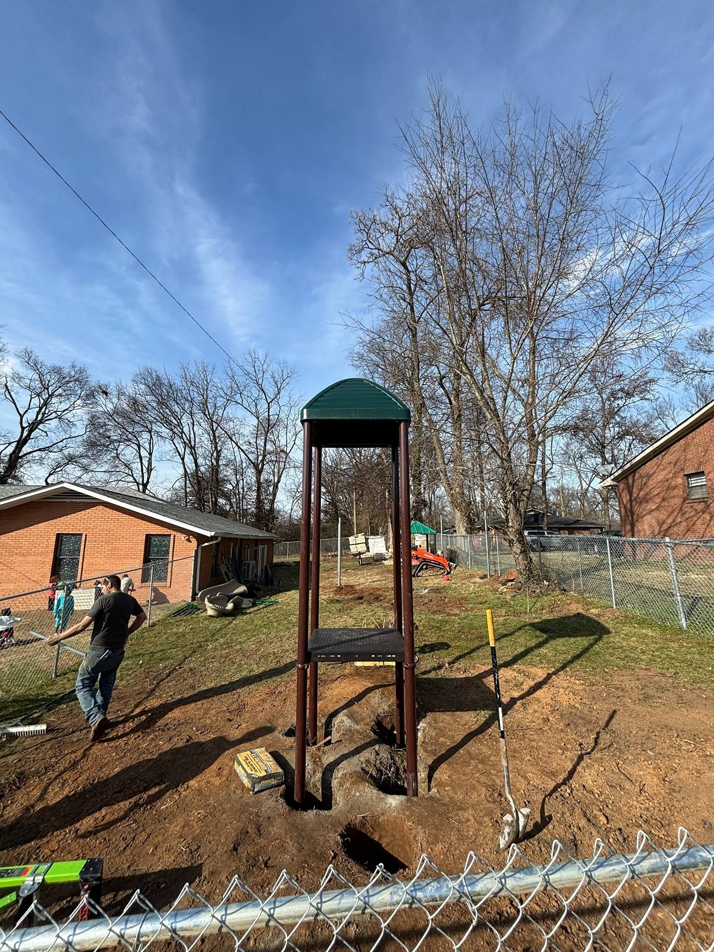 Small play tower with a green roof in a park; a person walks nearby on a sunny day.