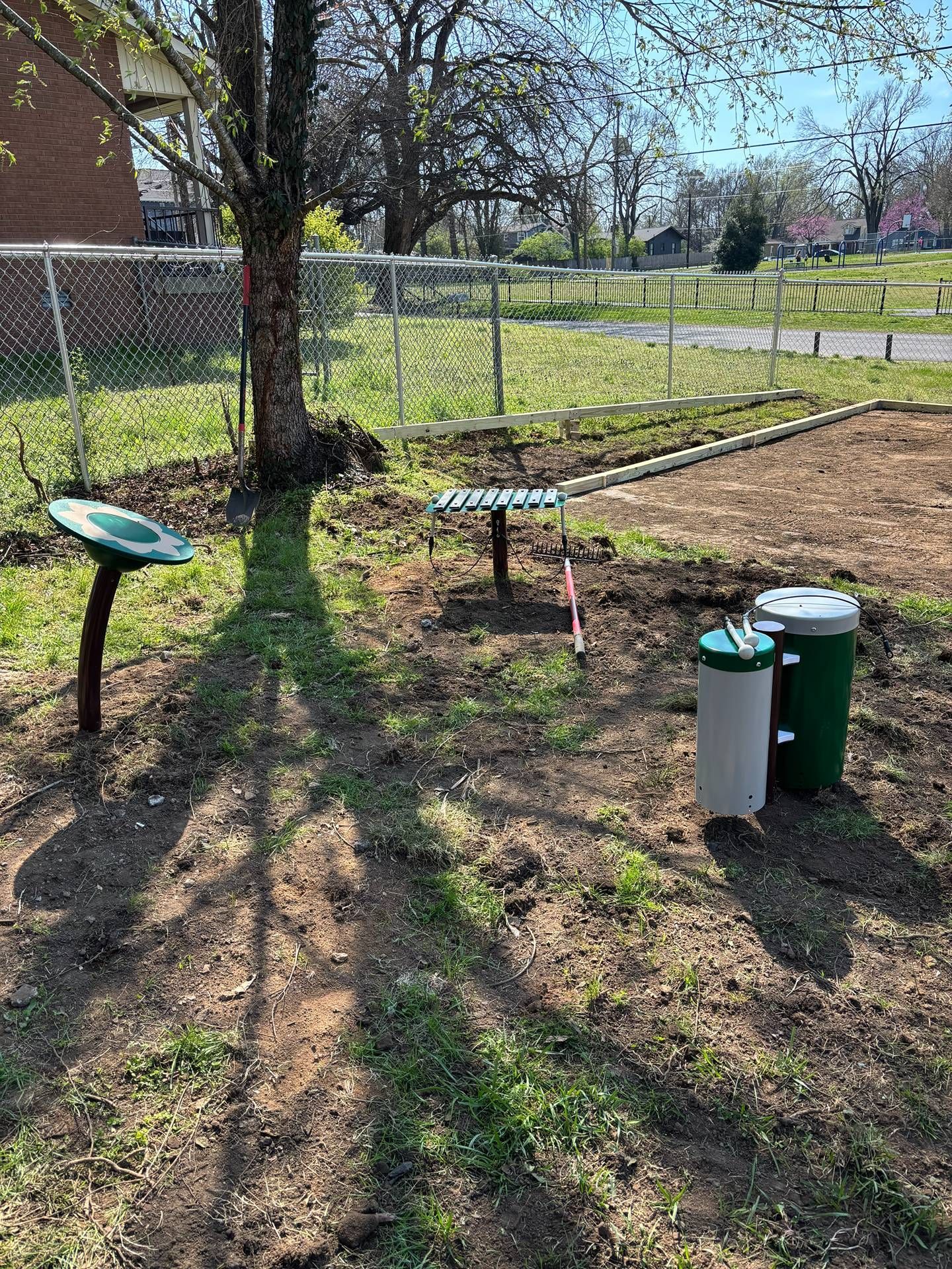 Outdoor exercise area with green fitness stations on grass beside a chain-link fence and trees.