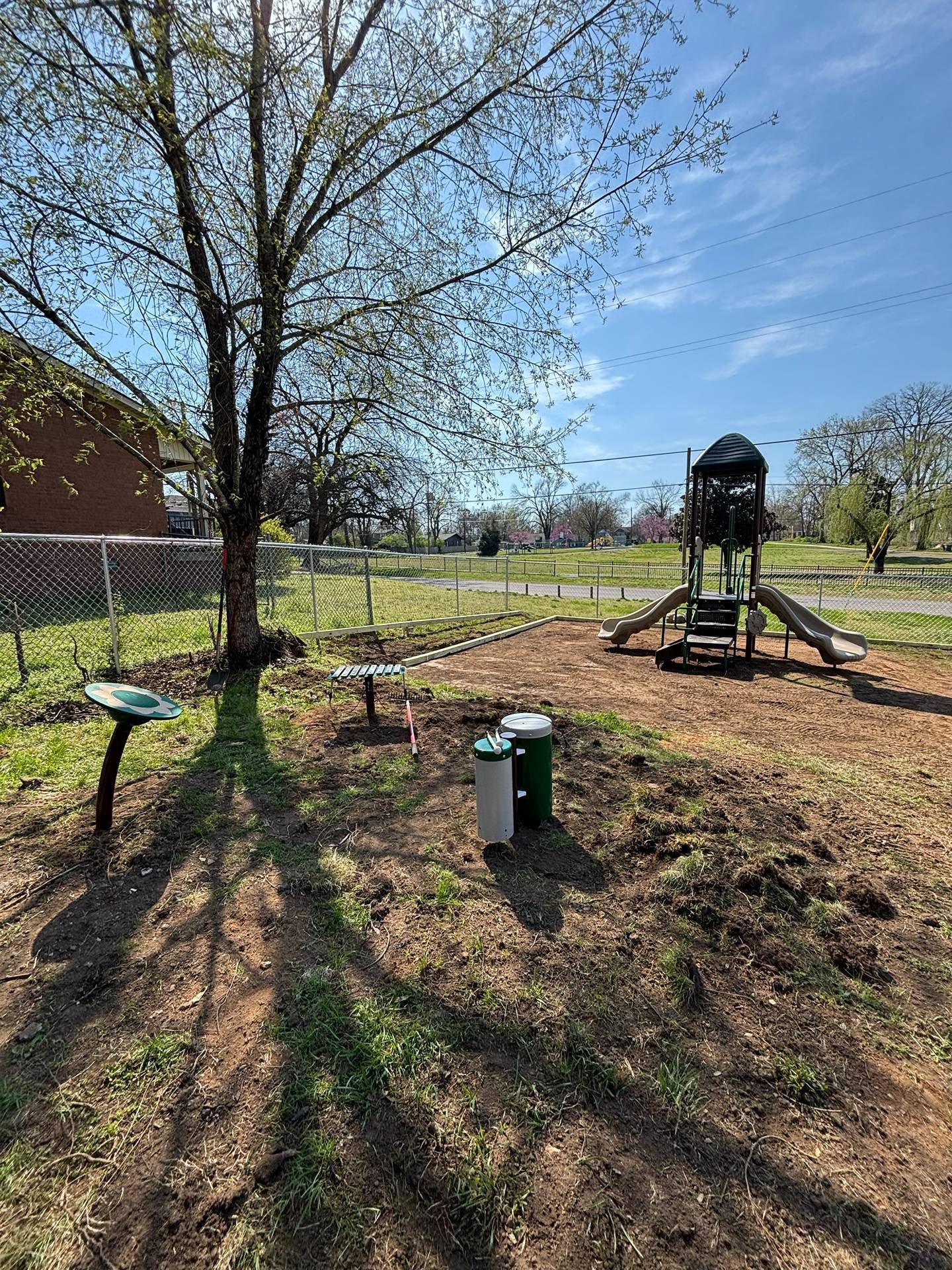 Sunny backyard with a large tree, a raised play structure, and scattered yard tools on bare ground.