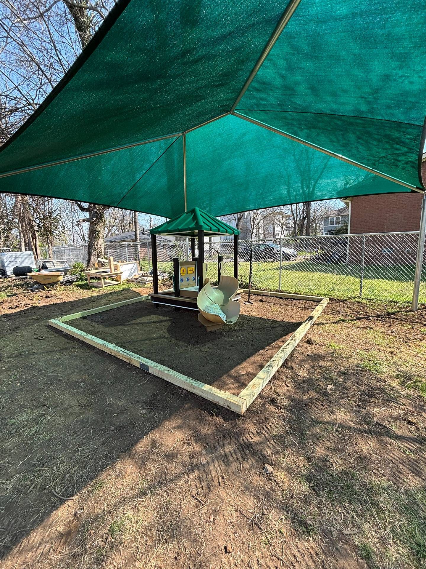 Shaded sandbox with green canopy, toy chairs, and a small playhouse in a fenced outdoor play area