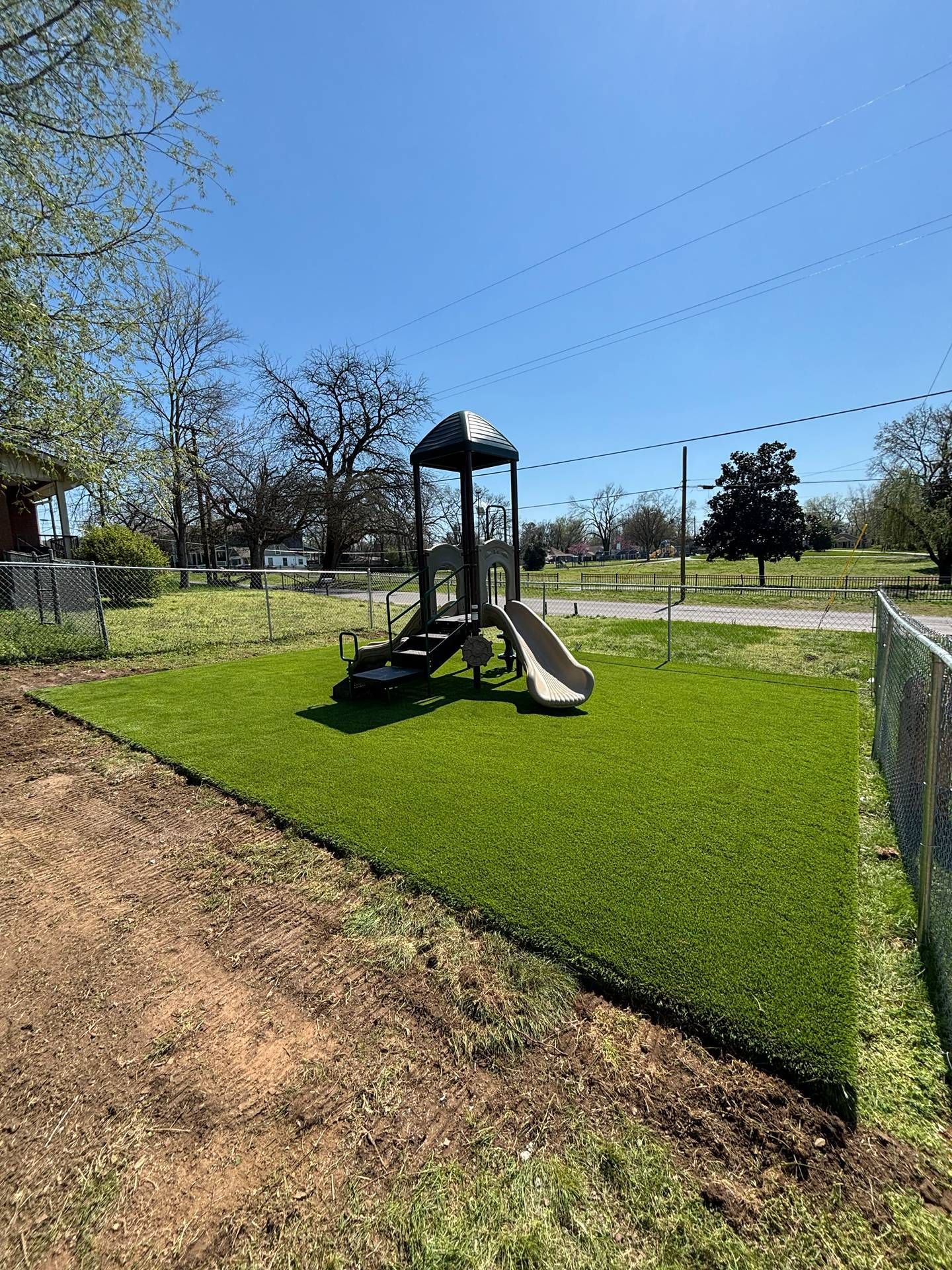 Small playground with slide and climbing tower on bright green grass beside a dirt path and chain-link fence.