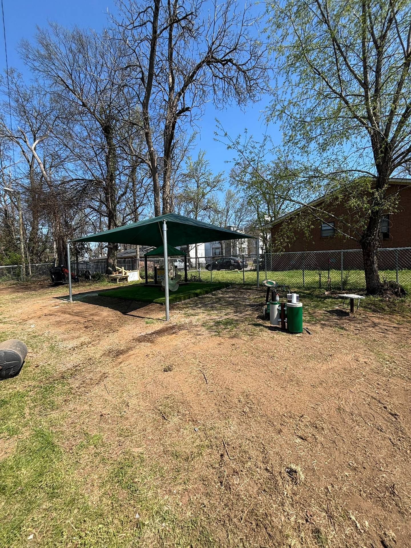 Park picnic shelter with a green roof, trees, and trash bins on a grassy slope.